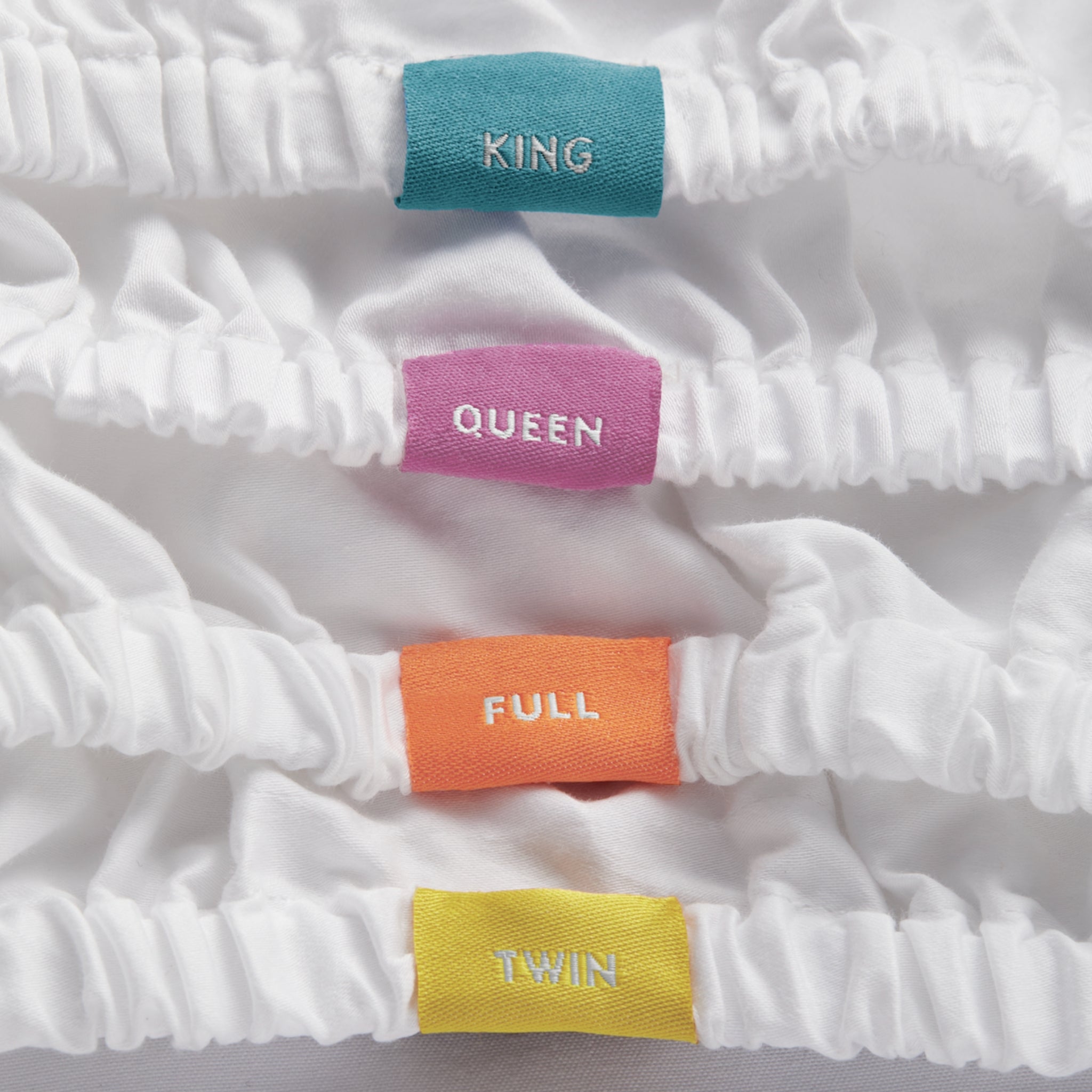 close up of tags on fitted sheets. Blue tag with the word King, pink tag with the word queen, orange tag with the word full, yellow tag with the word twin