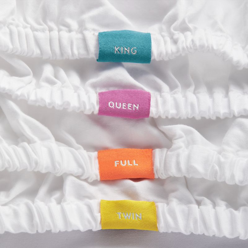 close up of tags on fitted sheets. Blue tag with the word King, pink tag with the word queen, orange tag with the word full, yellow tag with the word twin